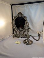 Photo showing silver plated ornate swivel mirror with cherub decorations alongside a three-arm candelabra on a white background.
