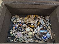 Overhead view of assorted costume jewelry densely arranged in mirror jewelry box, showing mixed bracelets, necklaces, earrings, rings, and pins.