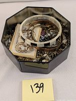 Jewelry collection in a black octagonal container showing mixed bracelets, pins, earrings, and chains in various metals and styles
