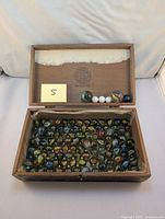 Wooden box open showing a large quantity of vintage ribbon glass marbles inside and larger marbles placed in the lid compartment.
