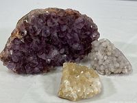 Three mineral specimens on a white surface: large purple amethyst cluster, smaller pale quartz/calcite cluster, and small white mineral cluster.