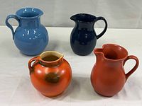 Group of four pottery pitchers on white surface: light blue glazed terra cotta marked Made in Italy with minor chips, dark blue unmarked, salmon marked Chantal, orange and brown hand-formed pitcher.