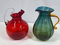 Both pitchers together showing sizes, colors, and overall shapes with amber handled pitcher on right and red pitcher with clear handle on left