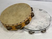 Photo showing two tambourines: one with natural skin drumhead, one with white coated drumhead with branding, side by side