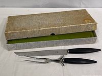 Full view of the two-piece carving set with original storage box. Knife and fork have black handles and polished metal blades/prongs.