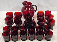 Full set of vintage ruby red glassware including pitcher, three sizes of tumblers, and cup and saucer sets arranged on white background.
