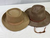 Two wool felt men's hats on white fabric, one light brown with ribbon band, one darker brown with leather band and bead.