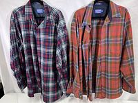 Two Pendleton wool lodge shirts hanging side by side, one in green, blue, and red plaid (tall), and the other in red, orange, and green plaid (likely the XXL long).