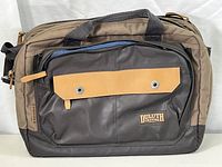 Front view of the convertible messenger bag showing brown and black fabric with zippered compartments and leather tab details.
