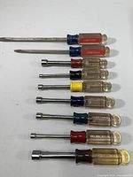 Full view of nine screwdrivers and nutdrivers with colored handles and metal shafts, arranged horizontally