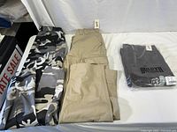 Three pairs of men's outdoor/work pants laid out flat: one black and white camo Boulder Creek pair and two tan Duluth pairs.