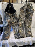 Full view of two Wolf Mountain insulated hunting pants hanging side by side, showing camo pattern and suspenders.