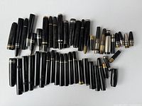 Top view of an assortment of black fountain pen parts including caps and barrels spread on a white surface, showing a variety of sizes and styles.