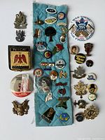 A collection of 34 various vintage pins on a blue cloth and a white surface, showing different designs including military, sports, and commemorative pins