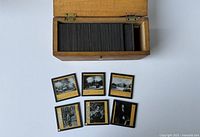 Wooden box containing many black bordered glass slides with yellowed backing. Six slides displayed showing monochrome historical imagery.