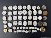 Overview of approximately 50 vintage pocket watch dials laid out on a black surface showing a range of sizes and conditions, some attached to movements.