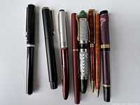 Top view of nine pens including seven fountain pens and two ballpoint pens displayed in a line, showing various colors and finishes