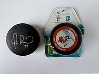 Photo showing two hockey pucks: one autographed black puck and another sealed commemorative Vancouver 2010 Olympics puck