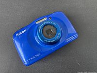 Front and top view of blue Nikon Coolpix point and shoot digital camera showing lens, flash, and markings indicating waterproof and shockproof capabilities.