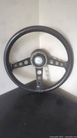 Full view of black leather wrapped 3 spoke steering wheel with circular holes in spokes, mounted against a corner wall.
