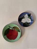 Two small vintage enamel plates side by side, showing one with white trillium on blue and one with red apple on green background.