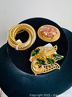 Five assorted gold tone vintage brooches arranged on black fabric - including a brooch with green rhinestones, a triple loop circular brooch, and oval-shaped brooches