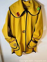 Full view of the yellow wool jacket hanging showing shape, collar, pockets and ceramic buttons with colorful stitching details.
