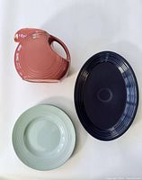 Fiesta Ware Dusty Rose ceramic pitcher, Navy oval serving platter, Woods Ware pale green Beryl plate all displayed flat on a white surface.