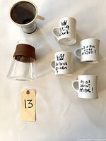 Top-down view of the pour over coffee maker components including stainless steel mesh filter and glass carafe with brown silicone grip, alongside four white mugs arranged in a cluster with their texts visible