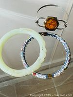 Top view of all three bracelets displayed on clear surface: pale green carved jade bracelet, blue and white enamel bracelet, and silver cuff with amber stone.