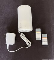 White cylindrical aroma diffuser with attached power adapter and two small bottles of essential oils labeled 'Yuzu' and 'Sweet Orange' placed on dark fabric background.