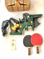 Vintage portable badminton set components including two metal clamps with nets, three shuttlecocks, two wooden paddles with red and black rubber surfaces, and a woven wicker basket.