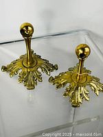 Two brass ornate bathroom accessories with leaf-pattern square bases and spherical finials shown on a clear surface