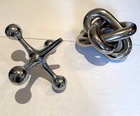Two polished metal sculptures: a jack-shaped piece and an intertwined knot piece on a white surface.