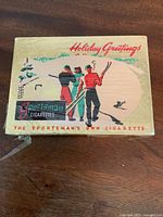 Front and top view of antique Sportsman Holiday Greetings cigarette tin showing illustrated winter sports scene with skiers and branding text.