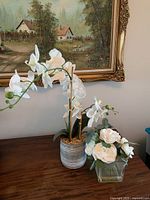 Two artificial flower arrangements displayed on a wooden surface in front of a framed painting. The first is white orchids in a metal bucket pot, and the second is white roses in a clear glass vase.