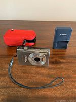 Compact Canon PowerShot SD750 camera, battery charger, and red carrying case arranged on a wooden surface