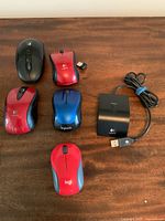 Five computer mice of various colors and styles including wireless models, and a Logitech docking station with USB cable arranged on wooden surface.