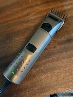 Metallic silver Conair men's electric shaver with black power switch, plugged into charger, resting on a wood surface.