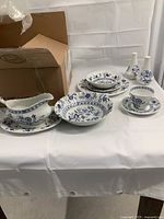 Photo showing assorted place setting pieces including gravy boat and serving platter with ornate blue floral pattern, placed on white cloth with open cardboard box in background.