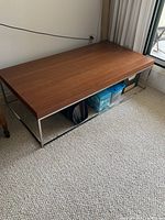 Full view of the rectangular wooden top coffee table on a carpeted floor, showing chrome base and space underneath with storage boxes.