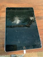 Front view of iPad showing large cracks on the glass and screen turned off. Device is plugged in via charging cable.