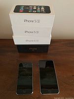 Photo showing four iPhone original boxes stacked; two iPhone 5S boxes, one iPhone 6 box, and one iPhone 3G box, with three iPhones placed in front, with one cracked screen visible on an iPhone 5S.