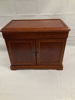 Closed view of the wooden jewelry box with reddish-brown finish and decorative front double doors with metal handles.