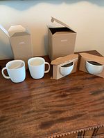 Four white Nespresso coffee cups displayed on a wooden surface with their original boxes. Two larger mugs and two smaller espresso cups.