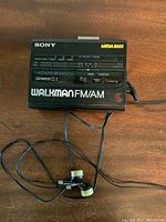 Front view of black Sony Walkman with FM/AM radio dial and Mega Bass logo, wired earbuds attached, on wooden surface.