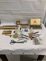 Overview photo of all items arranged on a white cloth background showing rulers, scissors in variety of sizes, a small clock, small boxes, and metal tray.