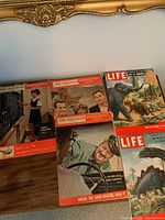 Lot of five vintage magazines displayed on wooden surface with ornate gold frame above them. Magazines include three Weekend (The Telegram) and two LIFE issues with visible covers showing different themes.