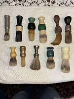 Full set of 11 antique and vintage men's shaving brushes displayed on a white towel. Variety of handle colors and materials visible including metal, wood, plastic, and resin.