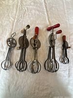 Photo showing all five antique hand mixers together on a white fabric background, displaying varying sizes and handle colors.